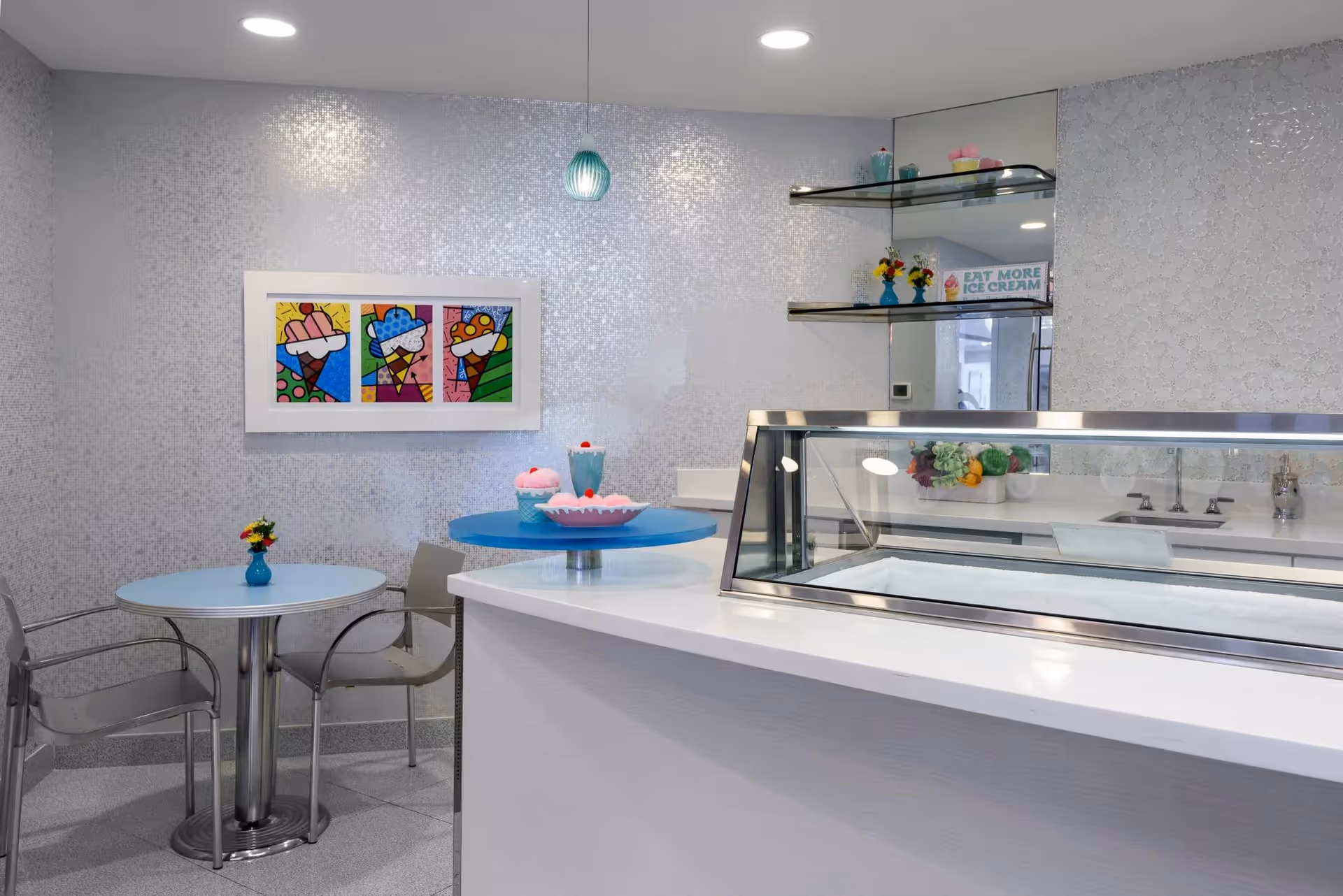 Interior view of a small ice cream or dessert serving area with a white counter and glass display case. Behind the counter are shelves with decorative items and a sign that says 'EAT MORE ICE CREAM'. To the left, there is a small round table with two chairs and a small vase with flowers. On the wall above the table is colorful artwork featuring ice cream cones. The walls have a light mosaic tile pattern and the ceiling has recessed lighting.