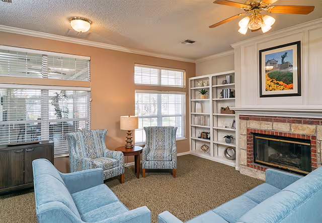 A cozy living room with two blue patterned armchairs and two blue sofas arranged around a small wooden side table with a lamp. The room features large windows with white blinds, a built-in white bookshelf with decorative items, and a brick fireplace with a framed picture above it. The ceiling has a light fixture and a ceiling fan with lights.