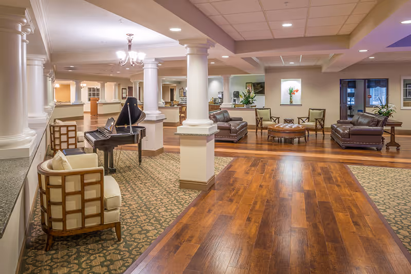 A spacious and well-lit assisted living facility common area with wooden flooring and carpeted sections. The room features a grand piano, several cushioned armchairs, leather sofas, a round ottoman, and decorative columns. There are flowers in vases placed on tables and wall niches, creating a welcoming and comfortable atmosphere.