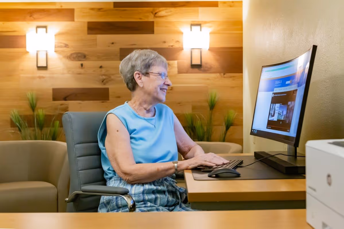 An elderly woman with short gray hair and glasses is sitting at a desk using a computer in a cozy room with wooden panel walls and soft lighting. She is smiling and appears engaged with the screen. There are two wall-mounted lights and some green plants in the background.