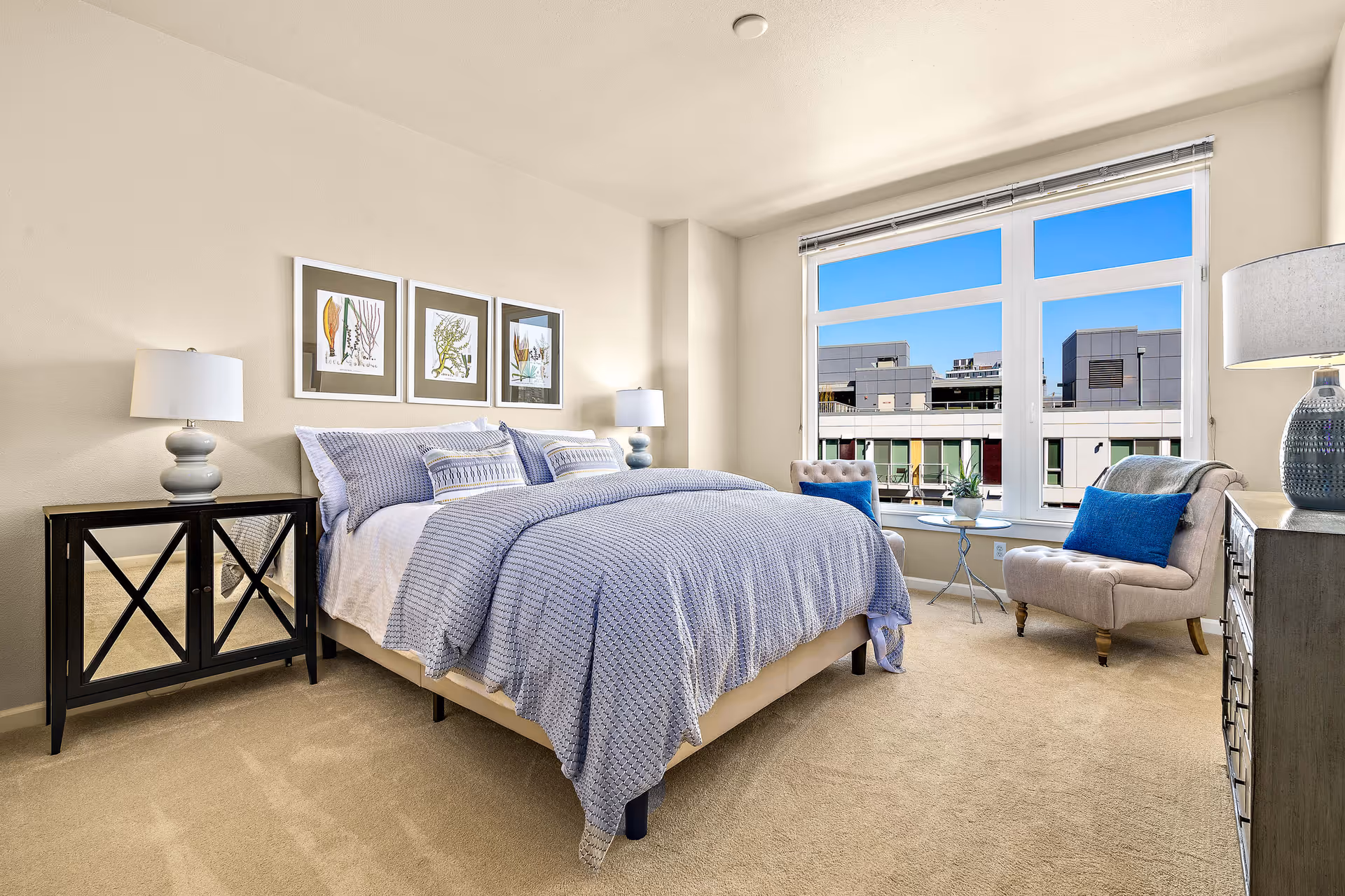A bright and modern bedroom with a large bed covered in a textured blue blanket and multiple pillows. On either side of the bed are matching bedside tables with lamps. Above the bed are three framed botanical prints. Near a large window with a view of nearby buildings, there are two beige upholstered chairs with blue cushions and a small round table between them. A dresser with a lamp is positioned on the right side of the room. The room has beige carpeting and light-colored walls.