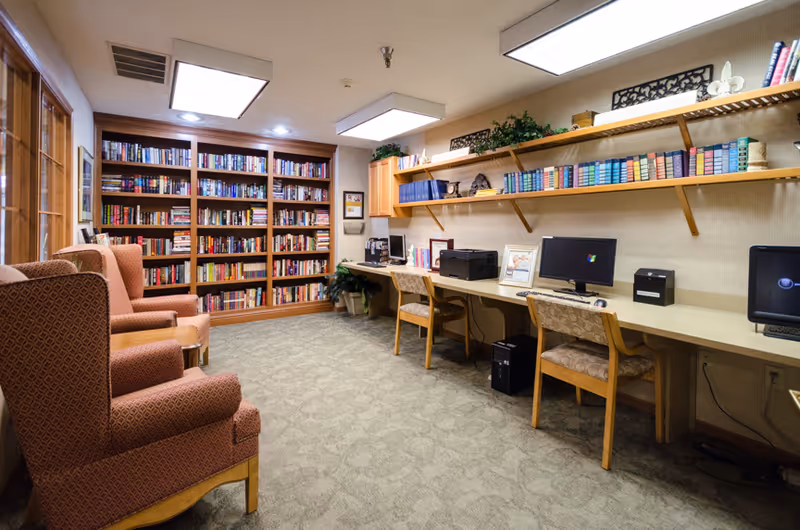A cozy library and computer room with bookshelves filled with books on the left wall, three upholstered armchairs in front of the shelves, and a long desk with three computer workstations and chairs on the right wall. Above the desk are wooden shelves holding books and decorative items. The room has carpeted flooring and soft lighting from ceiling fixtures.