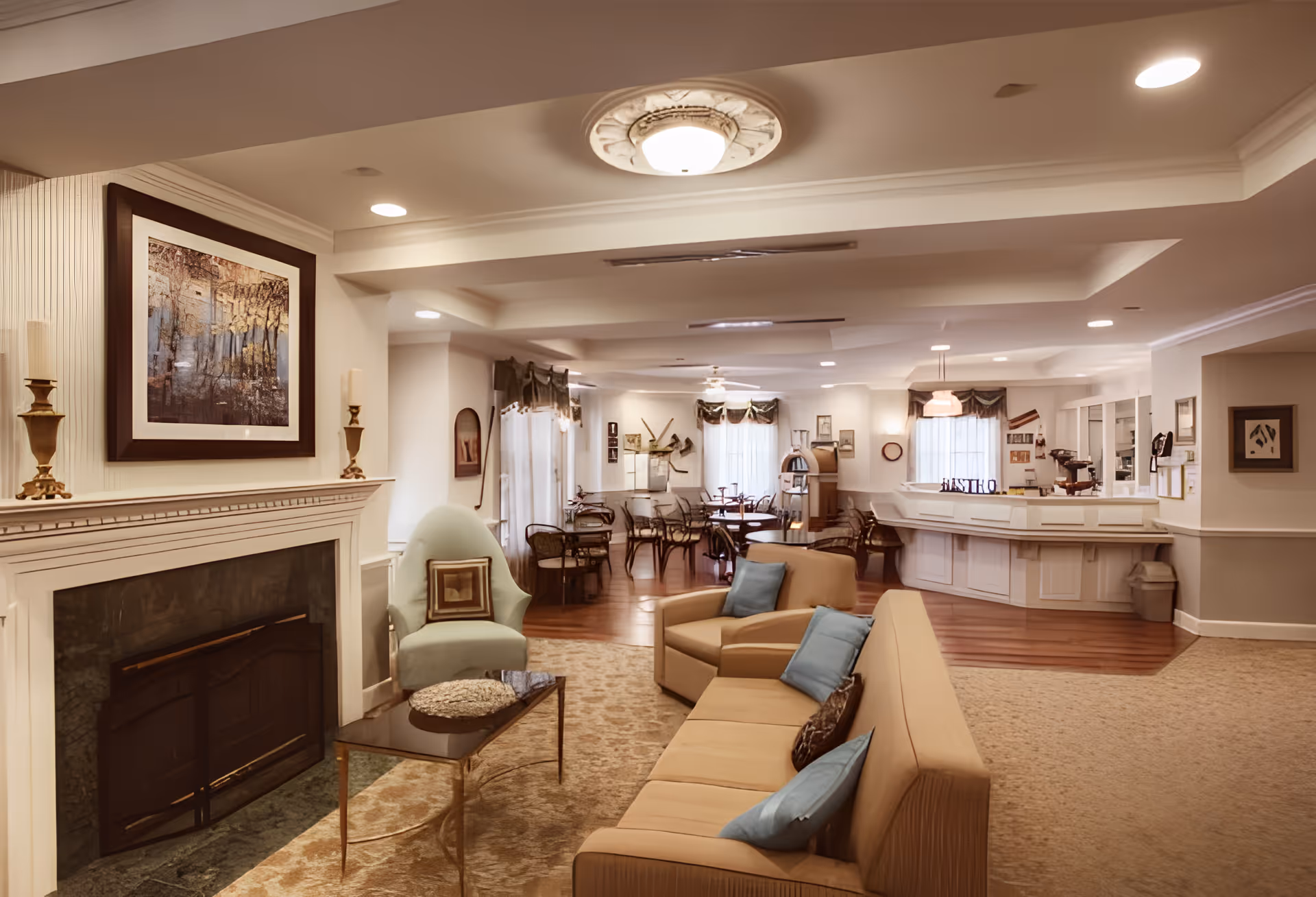A cozy senior living facility common area with a beige sofa adorned with blue and patterned pillows, a light green armchair, and a glass coffee table on a patterned rug. There is a fireplace with two candlesticks and a framed painting above it. In the background, there are several round tables with chairs, a counter area, and windows with curtains allowing natural light to fill the room.