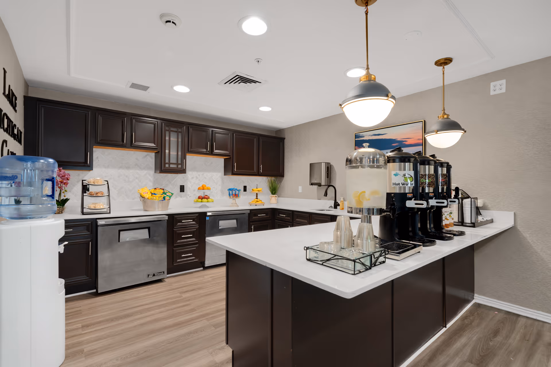 A modern kitchen area with dark wood cabinets, stainless steel dishwasher, and a white countertop island. The island has beverage dispensers including hot water and lemon water, along with plastic cups. The kitchen has wood flooring, pendant lights, and a water cooler on the left side.