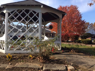 A white wooden gazebo on landscaped grounds with autumn foliage and buildings in the background.