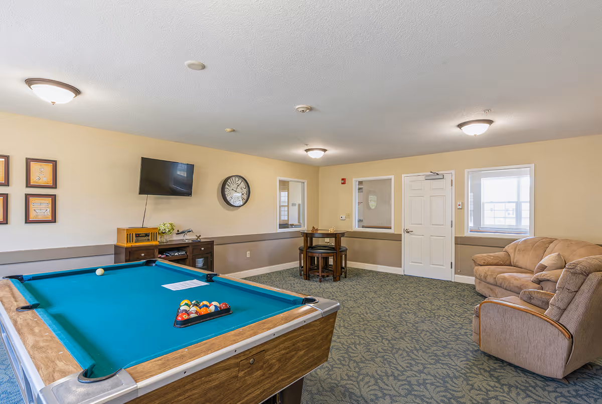 A recreational room in Allen Place Senior Living featuring a pool table with balls arranged for a game, a wall-mounted TV, a clock, a small table with chairs, a couch, and an armchair. The room has beige walls, carpeted floor, and ceiling lights.