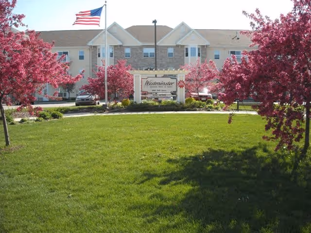 Front exterior view of Westminster Senior Housing building with an American flag on a flagpole, surrounded by blooming pink trees and a well-maintained green lawn.