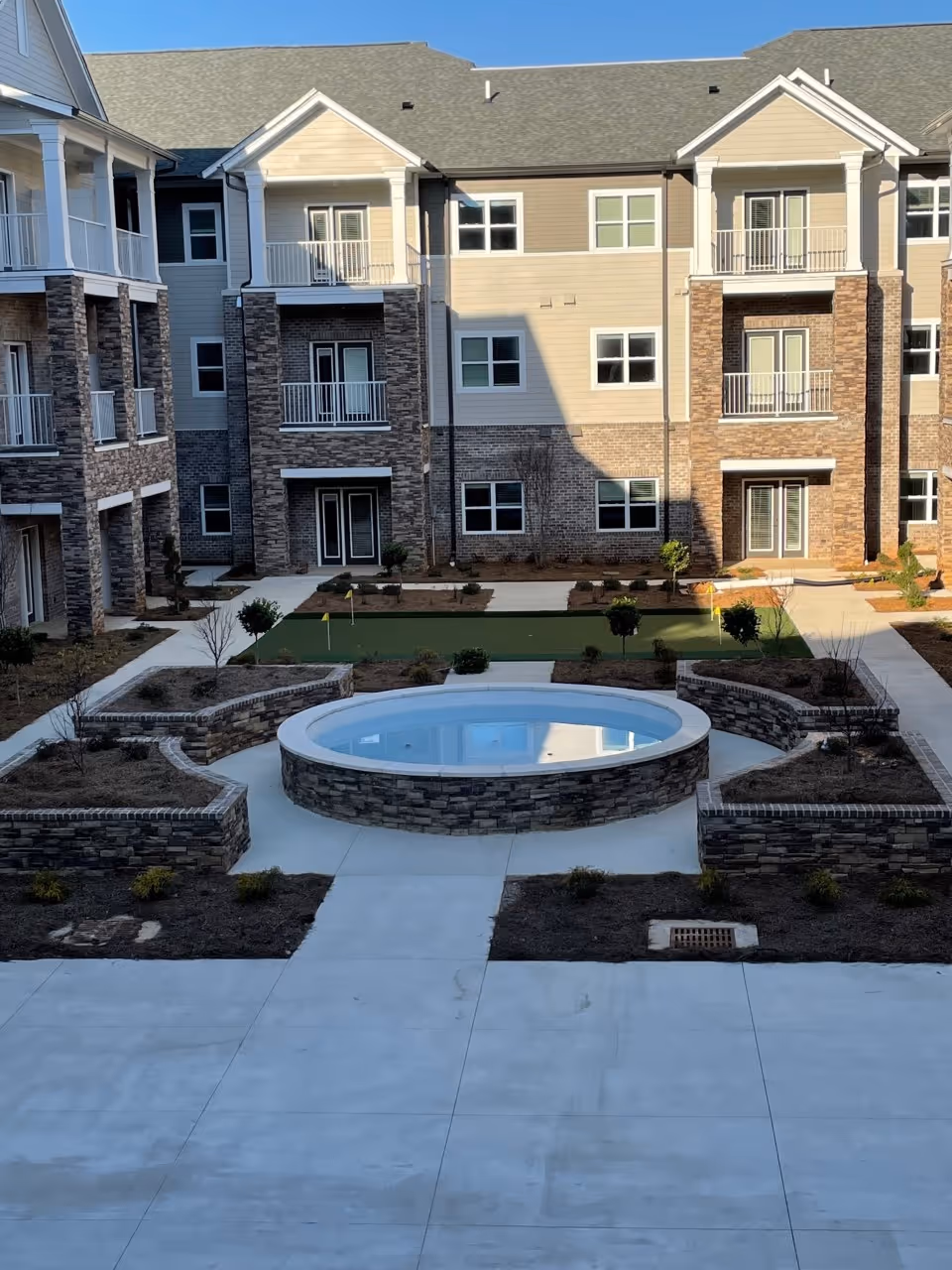 Outdoor courtyard area of a senior living facility with a circular water feature in the center, surrounded by stone planters and pathways. The courtyard is enclosed by a multi-story building with balconies and windows.