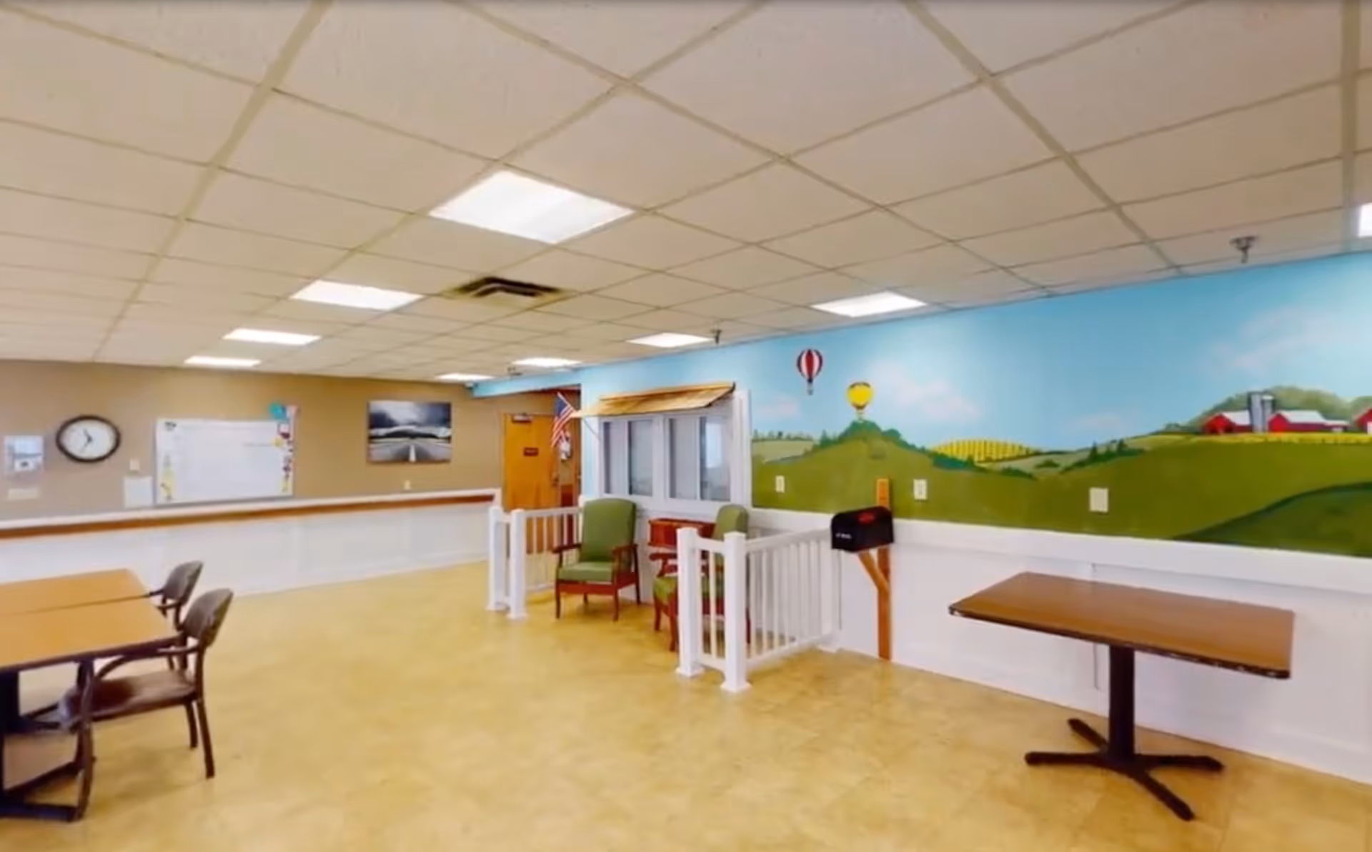 A spacious room with beige tiled flooring and a drop ceiling with fluorescent lights. There are several tables and chairs arranged around the room. On the right wall, there is a colorful mural depicting a countryside scene with green hills, hot air balloons, and a red barn. Two green chairs are placed near a small white fence and a mailbox attached to the wall. A clock and bulletin board are visible on the far left wall.