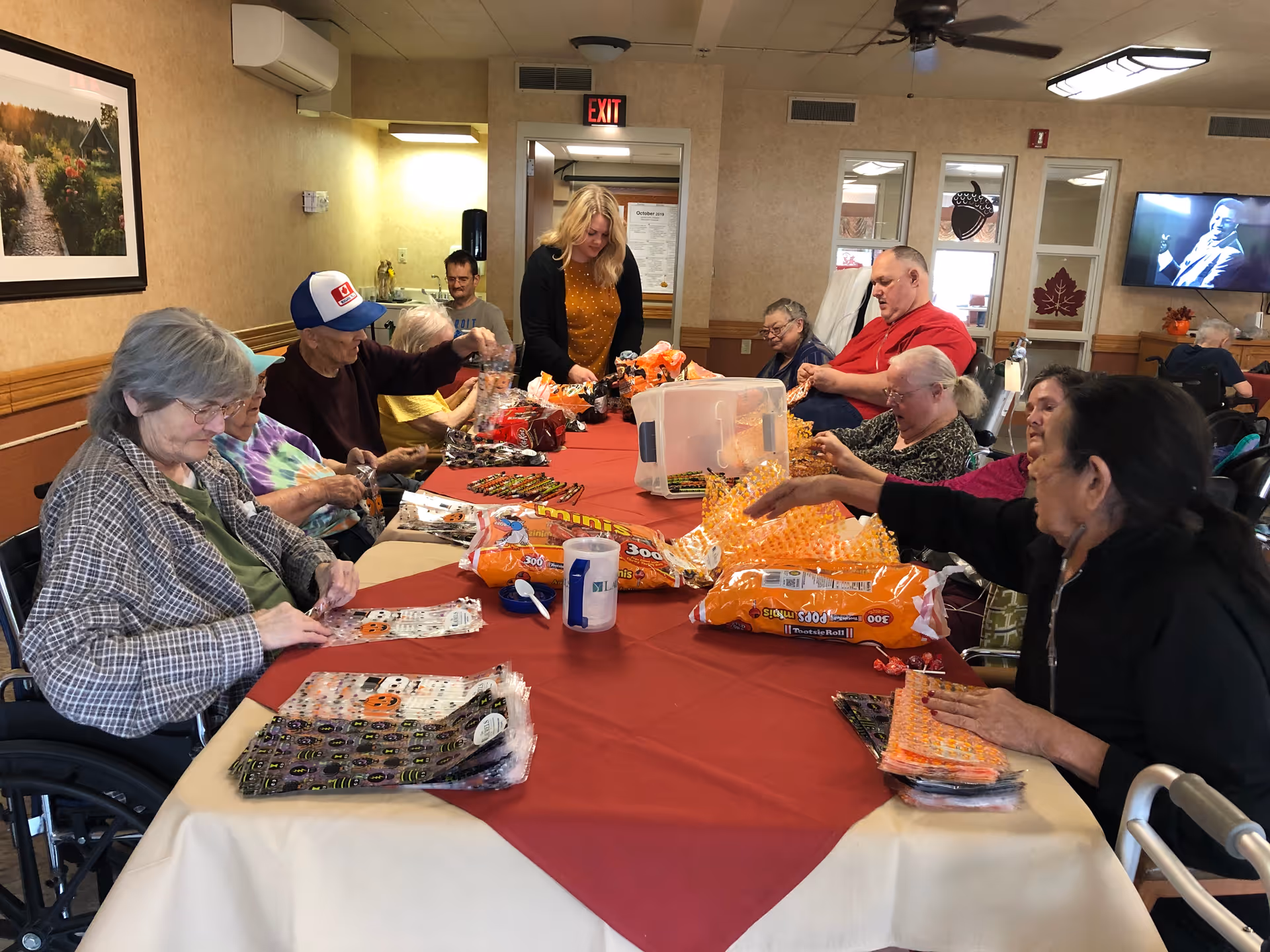 A group of elderly people sitting around a long table covered with a red tablecloth, engaging in a craft or activity involving orange and black materials. A younger woman stands at the far end of the table assisting them. The room has beige walls, framed pictures, and a television mounted on the wall.