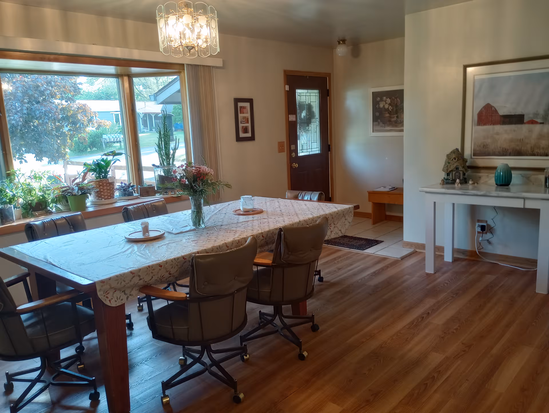 A dining room with a large rectangular table covered with a floral tablecloth and surrounded by six cushioned chairs on wheels. The table has a vase with flowers and some plates. There is a large window with several potted plants on the windowsill, letting in natural light. The room has wooden flooring, a chandelier hanging from the ceiling, a wooden door with a glass panel, and a white console table with decorative items and a framed picture on the wall.