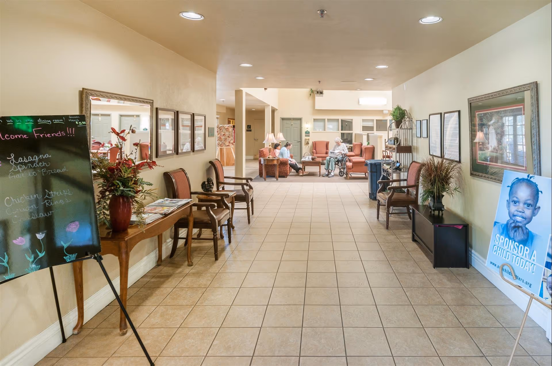 A wide hallway in an assisted living facility with tiled floors and beige walls. On the left side, there is a blackboard with a menu, a wooden table with a flower vase, and chairs. On the right side, there are framed pictures, a plant on a small cabinet, and a poster encouraging child sponsorship. At the end of the hallway, three elderly people are seated and engaged in conversation in a lounge area with armchairs and lamps.