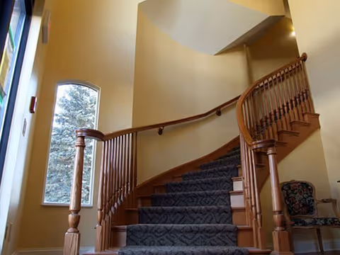 Curved wooden staircase with carpeted steps inside a building, featuring a large arched window on the left side and a floral upholstered chair on the right.