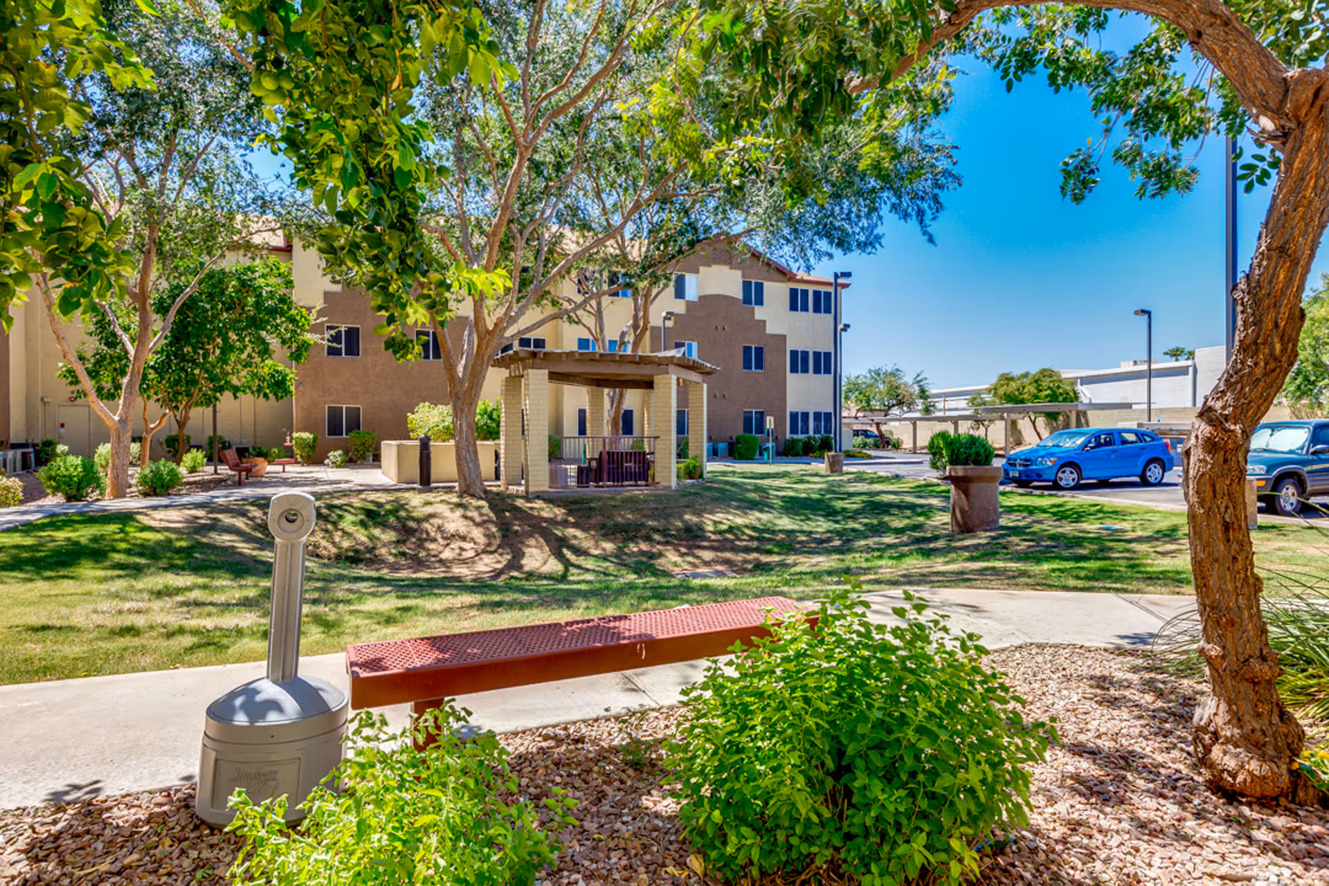 Outdoor garden area at Discovery Point Retirement Community with green grass, trees, bushes, a red bench, a covered seating area, and a parking lot with cars in the background under a clear blue sky.