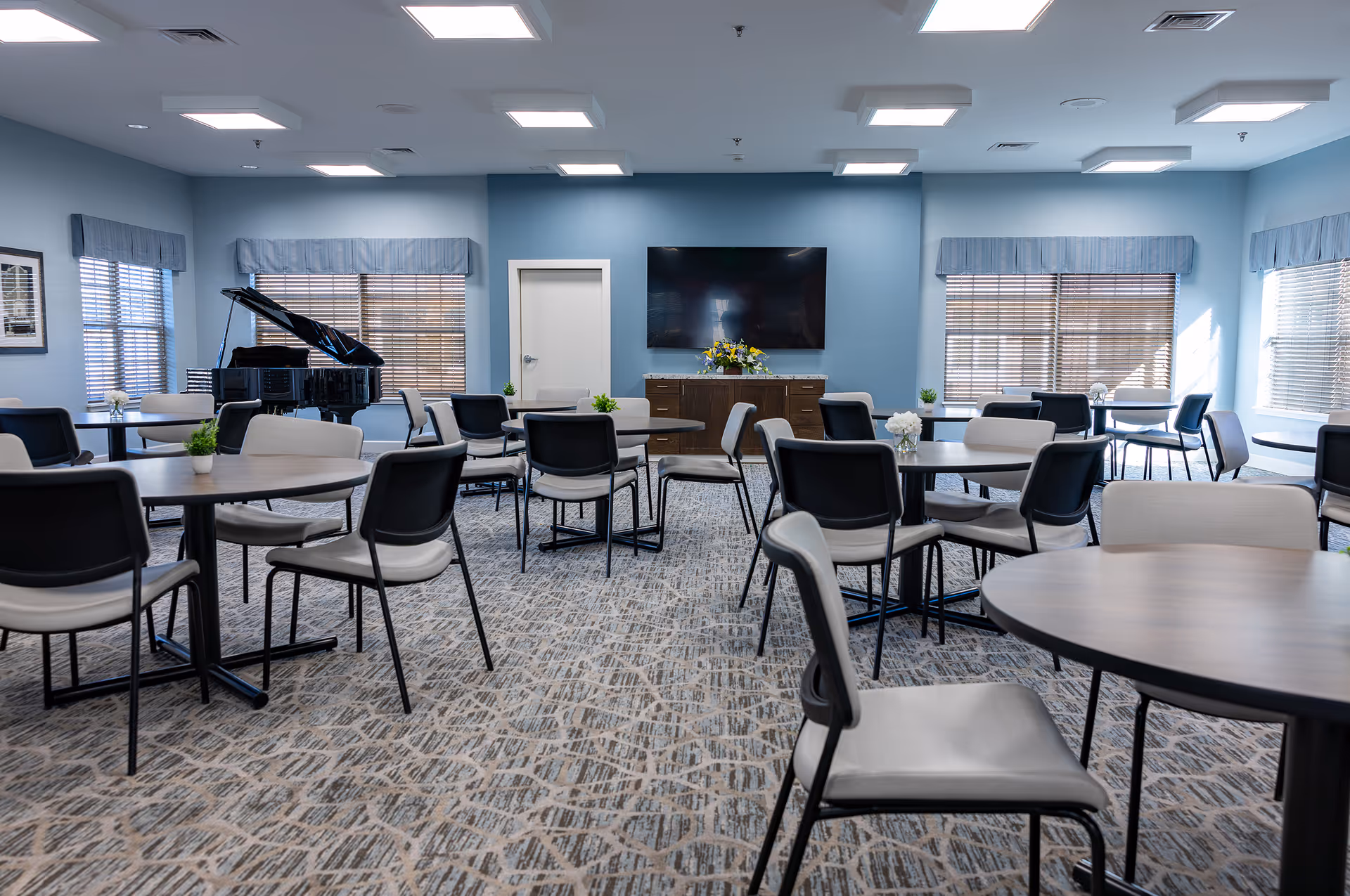 Spacious light-blue common room with round tables and chairs, a grand piano on the left and a wall-mounted TV at the center.