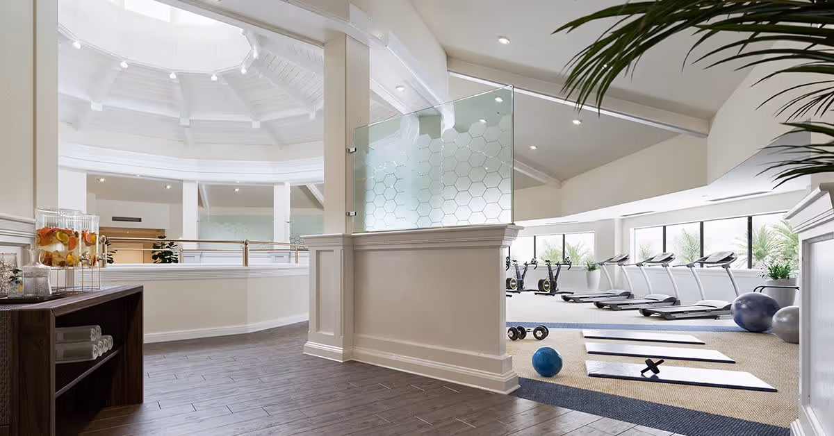 Bright and spacious senior living fitness room with several treadmills, exercise bikes, yoga mats, dumbbells, and exercise balls. The room features large windows letting in natural light, a high ceiling with white beams, and a refreshment station with infused water and towels on a wooden shelf.