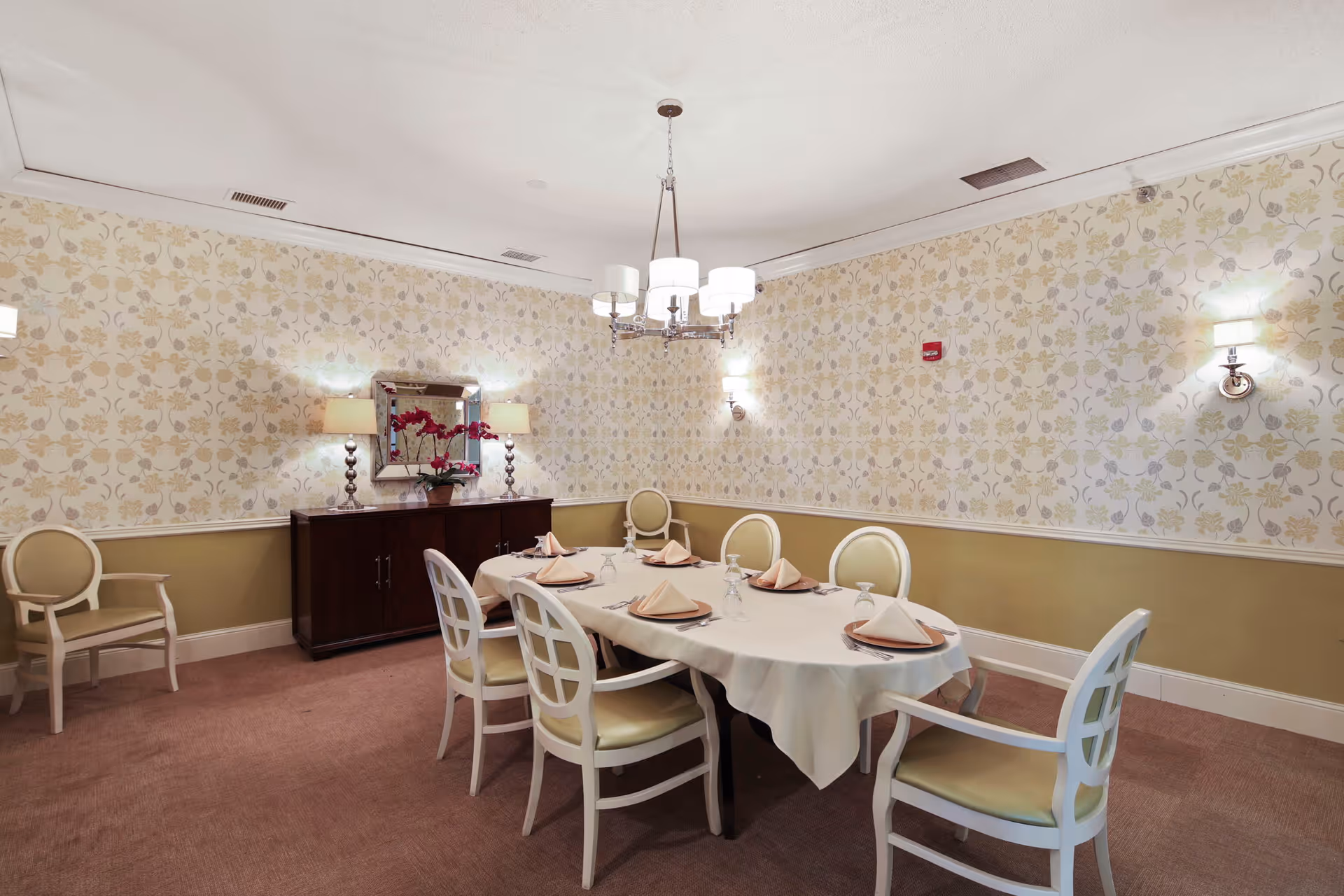 Small formal dining room with a table set for six, upholstered chairs, chandelier, sideboard and floral wallpaper.