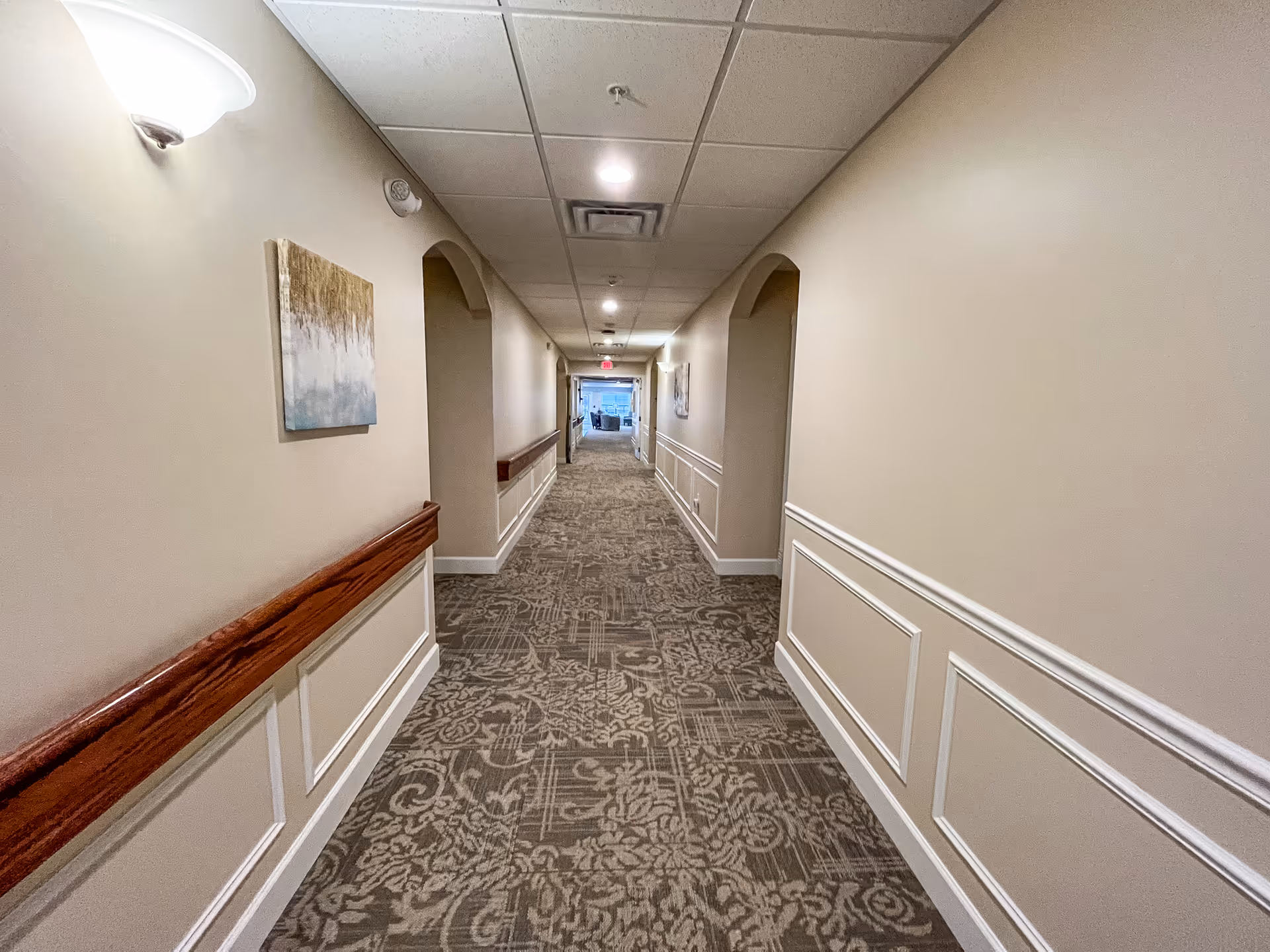 A long, well-lit hallway in an assisted living facility with beige walls, decorative white wainscoting, patterned carpet, and wooden handrails on both sides. There are wall-mounted lights and framed artwork along the corridor, with an exit sign and a seating area visible at the far end.