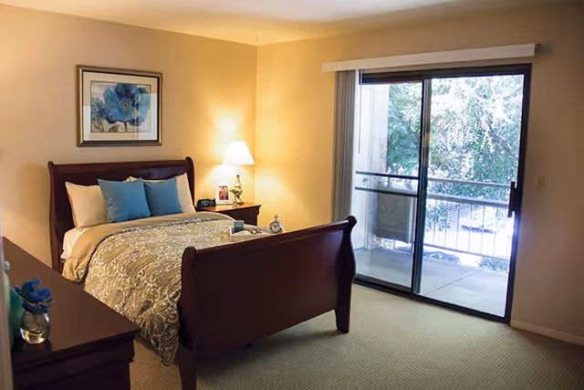 A cozy bedroom with a wooden sleigh bed made up with patterned bedding and blue accent pillows. There is a nightstand with a lamp, picture frames, and a clock beside the bed. A framed floral artwork hangs on the wall above the bed. Sliding glass doors lead to a balcony with a view of trees outside.