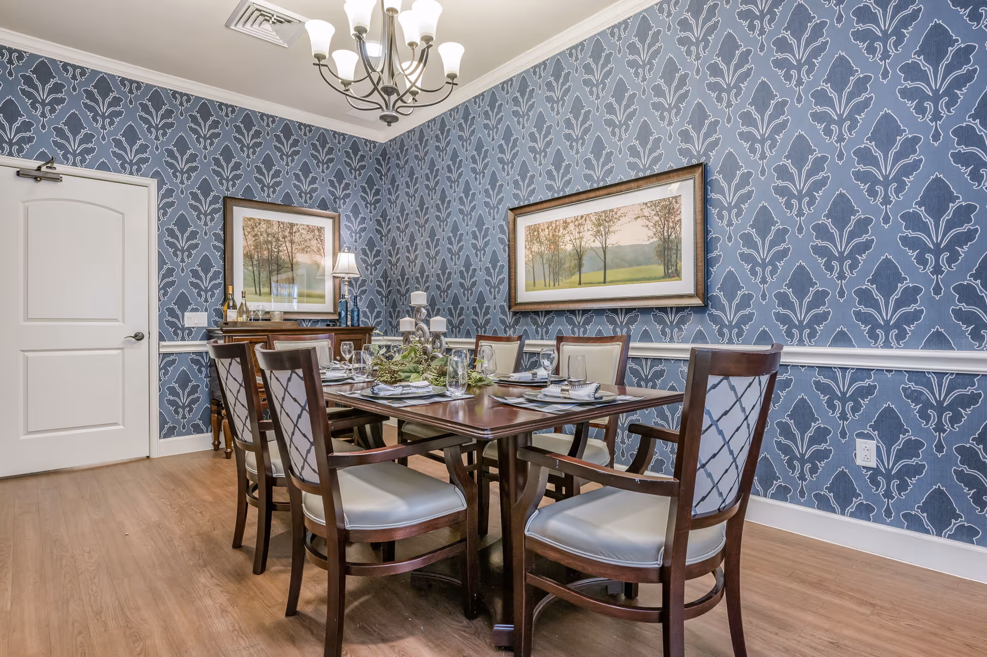 A dining room with a wooden table set for six people with plates, glasses, and napkins. The chairs have wooden frames with light-colored cushioned seats and backs. The walls are decorated with blue patterned wallpaper and two framed landscape paintings. A chandelier with multiple lights hangs from the ceiling, and a wooden sideboard with a lamp and decorative items is against the wall.