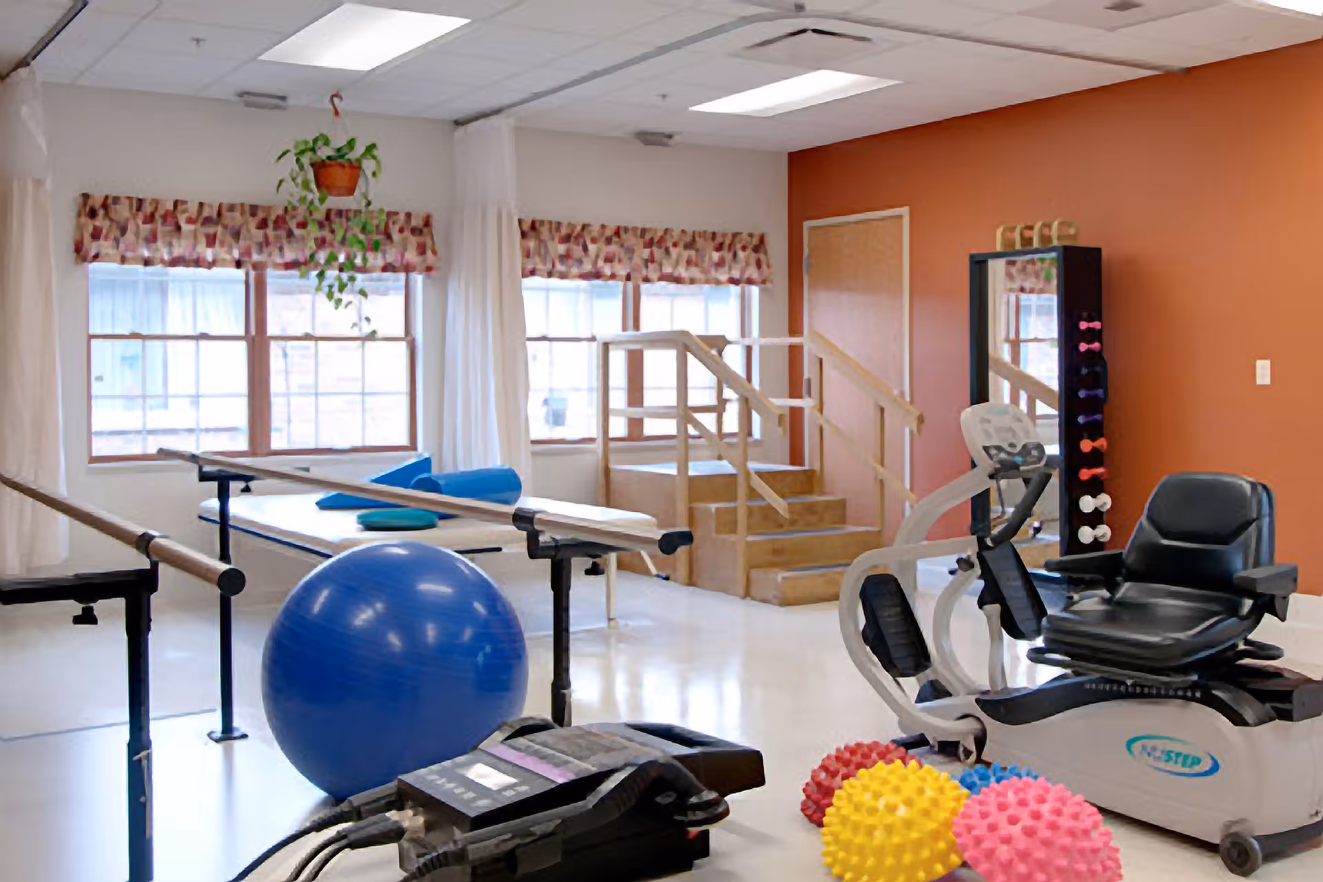 Physical therapy room with exercise equipment including a blue exercise ball, parallel bars, a recumbent exercise bike, colorful spiky massage balls, a padded therapy table, and wooden stairs with handrails. The room has large windows with patterned valances and a hanging plant.
