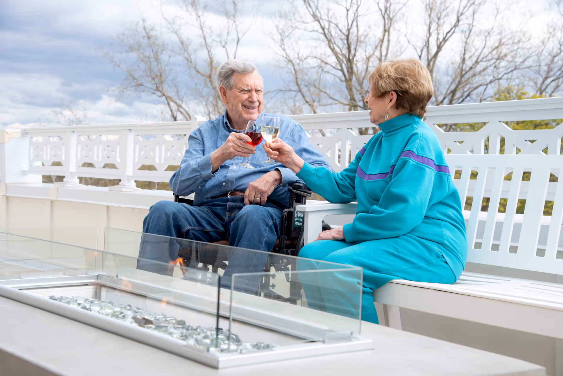 An elderly man in a wheelchair and an elderly woman sitting on a white bench outdoors, clinking their wine glasses together in a toast. They are on a patio with a modern fire pit table in front of them and a white decorative railing behind them. The sky is cloudy and there are leafless trees in the background.