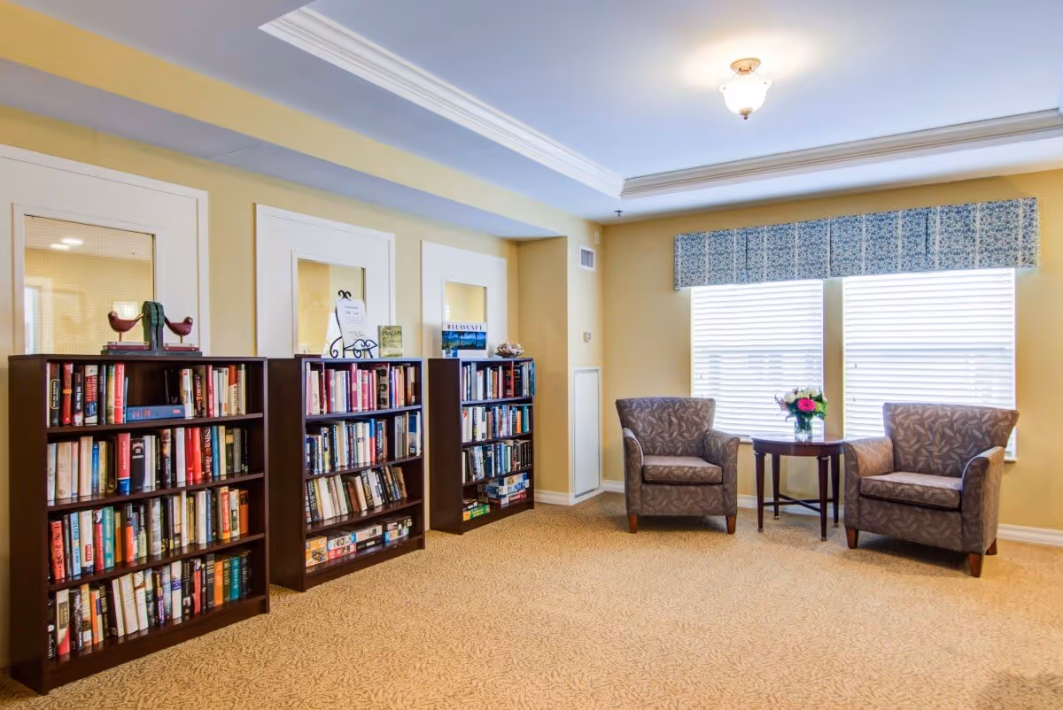 A cozy reading area in a senior living facility with three dark wood bookshelves filled with books against a yellow wall. Two patterned armchairs are placed near two large windows with blinds and a blue valance. A small round wooden table with a vase of flowers sits between the chairs. The room has beige carpet and a ceiling light fixture.