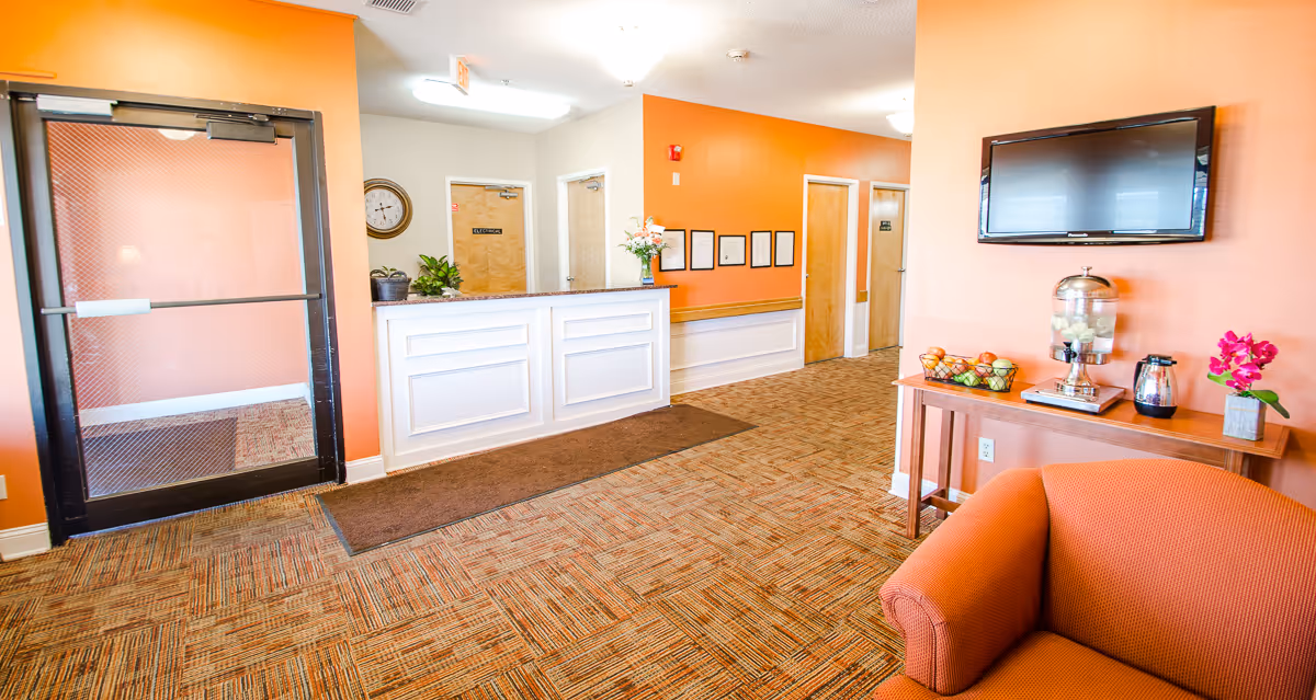 Reception area of a senior living facility with an orange accent wall, a white reception desk, a clock on the wall, and a seating area with an orange armchair. A wooden table holds a water dispenser, a coffee pot, a bowl of fruit, and a pink flower. A flat-screen TV is mounted on the wall above the table.