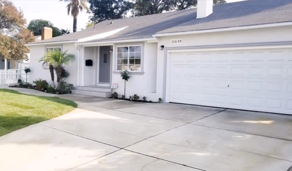 White single-story building with a two-car garage, front entrance, and palm trees nearby.