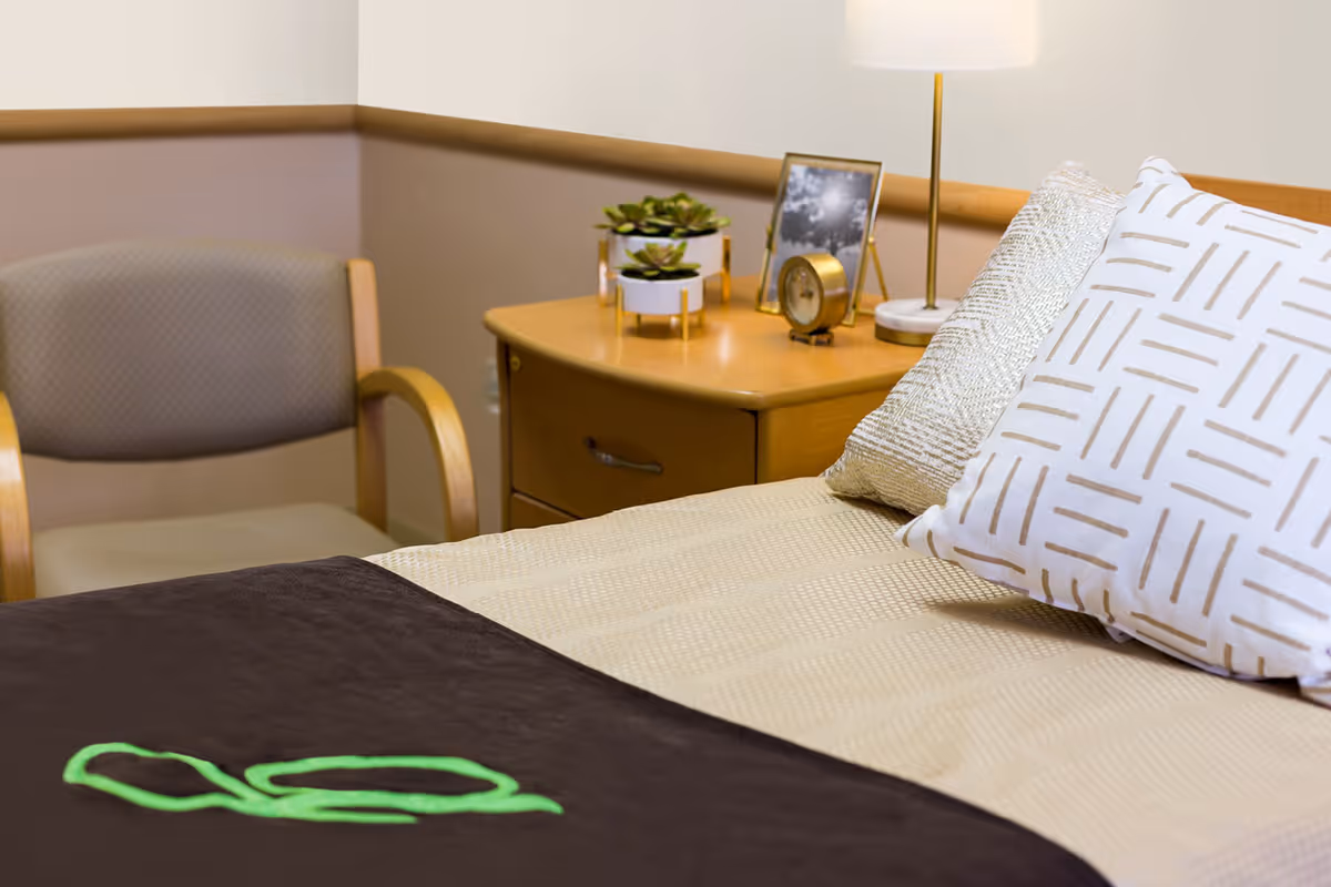 Corner of a furnished bedroom showing a made bed with decorative pillows, a nightstand with a lamp, clock and small plants, and a chair.