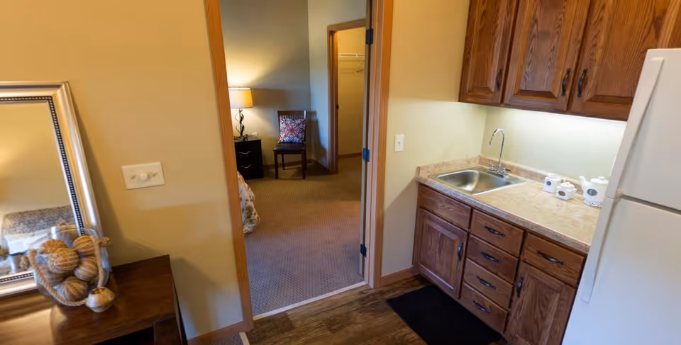 Small kitchenette with wooden cabinets, a sink and refrigerator opening into a bedroom where a chair and lamp are visible.