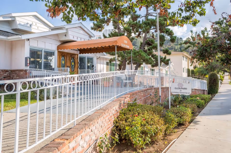 Exterior view of Glen Park at Glendale - Mariposa facility showing a white building with a brick base, a white metal fence, a sidewalk, and landscaping with bushes and trees. There is an orange awning above the entrance door with the text '1250 Glen Park'. A sign near the sidewalk reads 'Glen Park at Glendale Boynton St'.