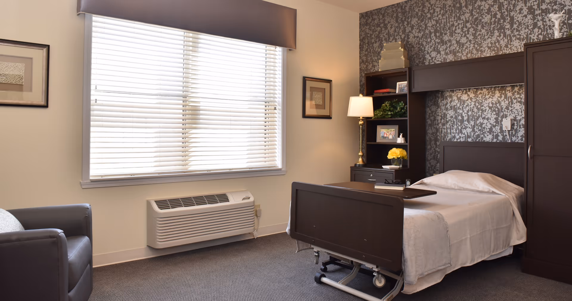 A neatly arranged bedroom in a senior living facility featuring a single hospital-style bed with white bedding, a dark wooden headboard and footboard, a bedside table with a lamp and decorative items, a dark armchair, and a large window with closed blinds allowing natural light to fill the room.
