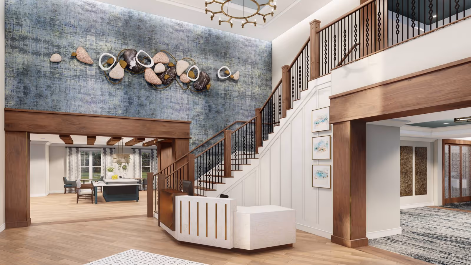 A spacious and elegant interior lobby area featuring a modern reception desk in front of a staircase with wooden handrails and black balusters. The wall behind the staircase has a textured blue wallpaper with decorative abstract wall art. To the left, there is an open dining area with a large island, chairs, and windows with curtains letting in natural light. The flooring is light wood, and the ceiling has a stylish chandelier.