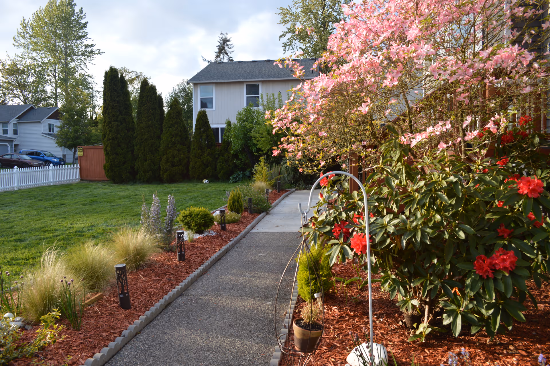 A landscaped outdoor pathway lined with various plants and flowers, including pink and red blossoms. The path is bordered by mulch and small garden lights, leading towards a residential building with trees and a grassy lawn on the left side.