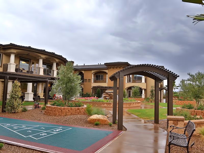 Outdoor courtyard area of a senior living facility with a shuffleboard court, a wooden pergola over a walkway, landscaped garden beds with trees and flowers, benches, and a two-story building with balconies in the background under a cloudy sky.