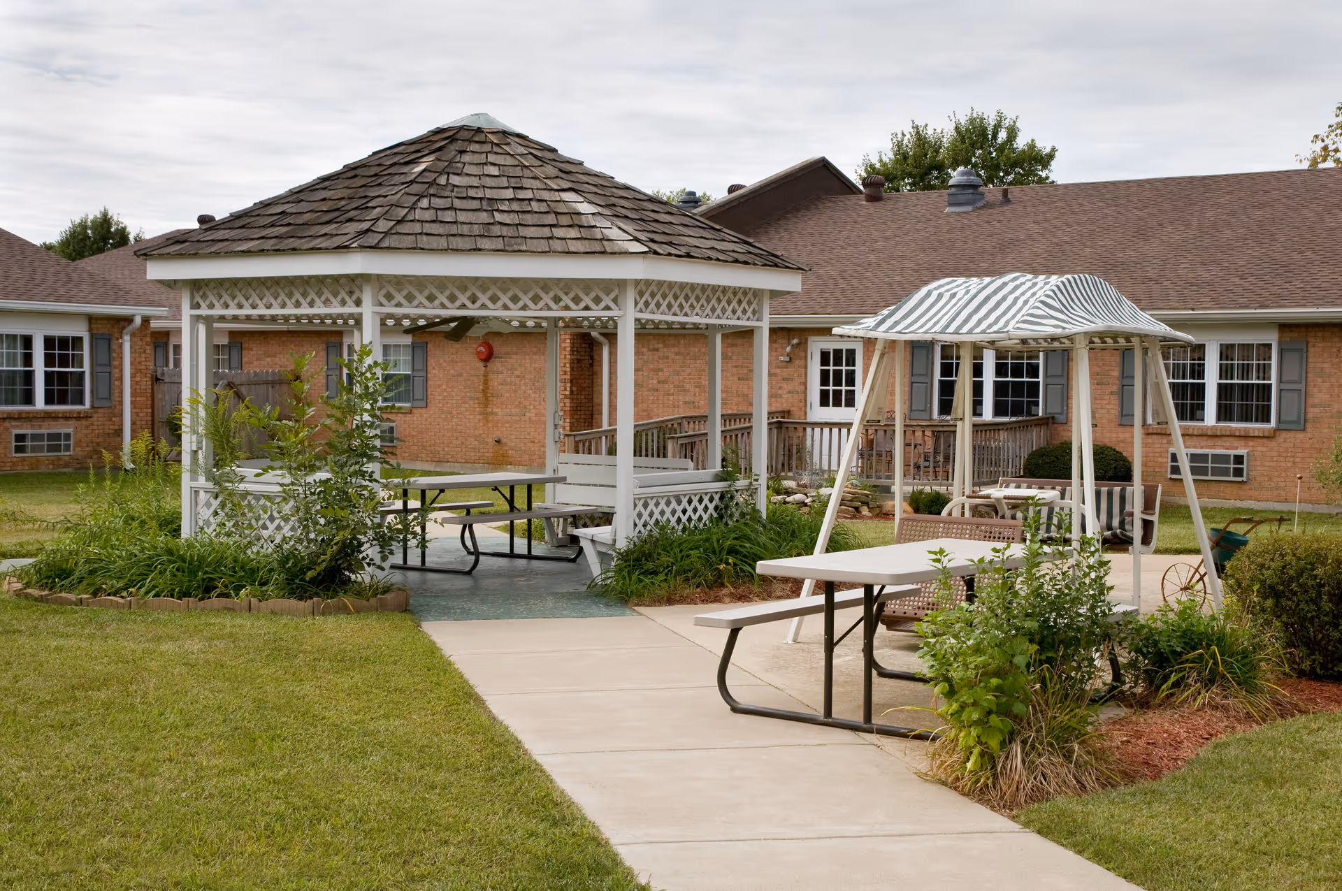Courtyard with a white gazebo, a striped canopy over a picnic table, and a brick care facility building in the background.
