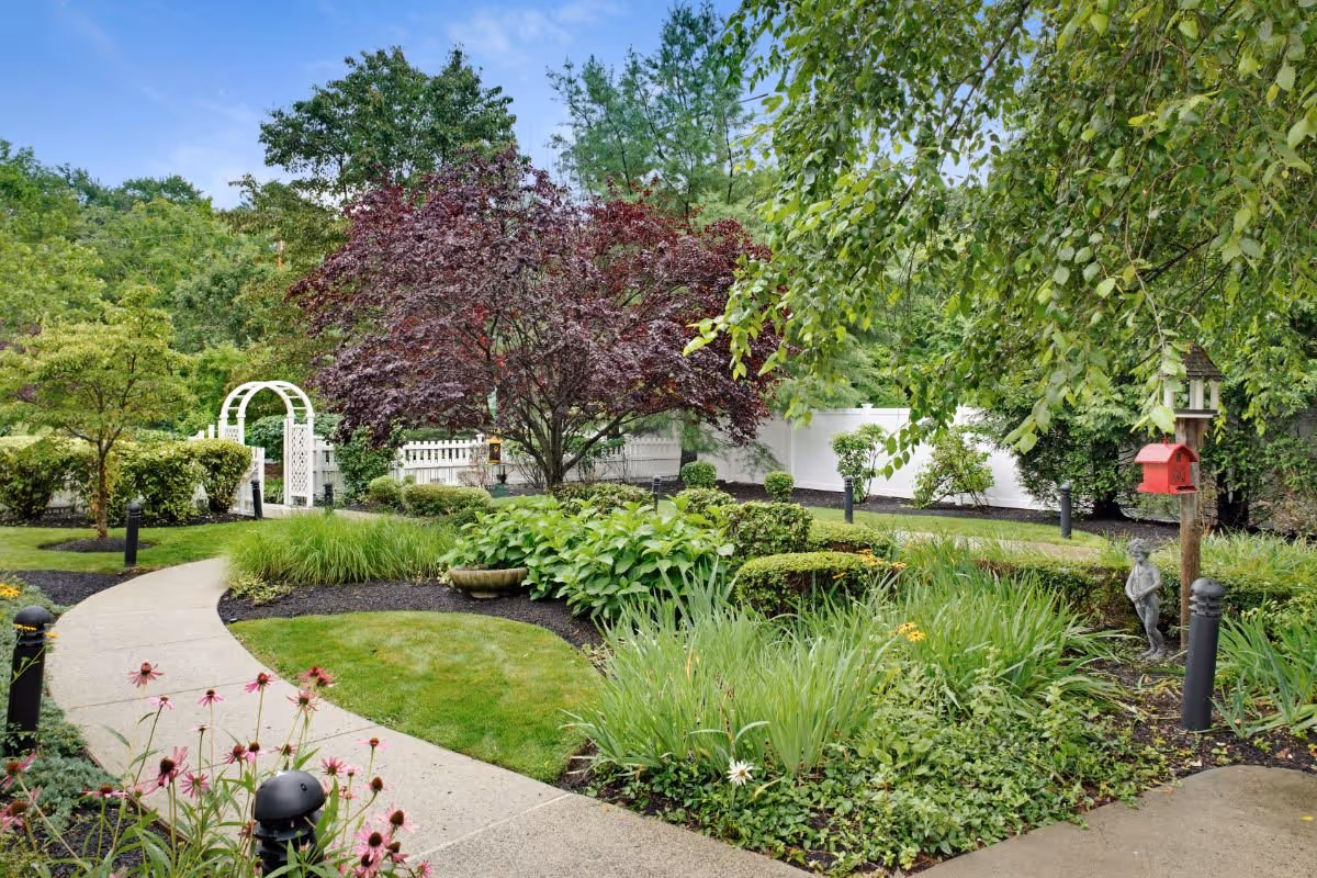 A well-maintained garden area with a curved concrete pathway surrounded by various green plants, bushes, and trees. There is a white wooden archway in the background, a red birdhouse mounted on a post, and a small statue of a child near the plants. The garden is enclosed by a white fence and has several black garden lights along the path.