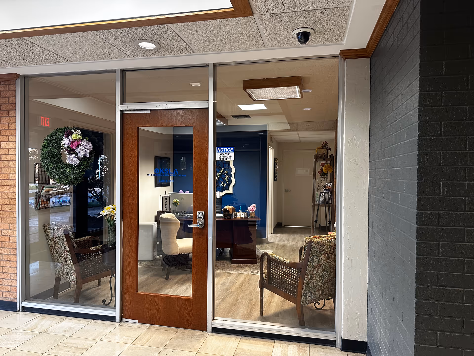 Glass-fronted reception office with a wooden door, seating, desk, and decorations inside a senior living facility.