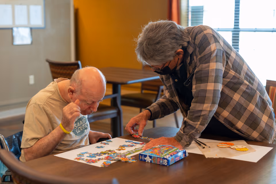 An elderly man and a caregiver working together on assembling a colorful jigsaw puzzle at a wooden table in a well-lit room with chairs and a window in the background.
