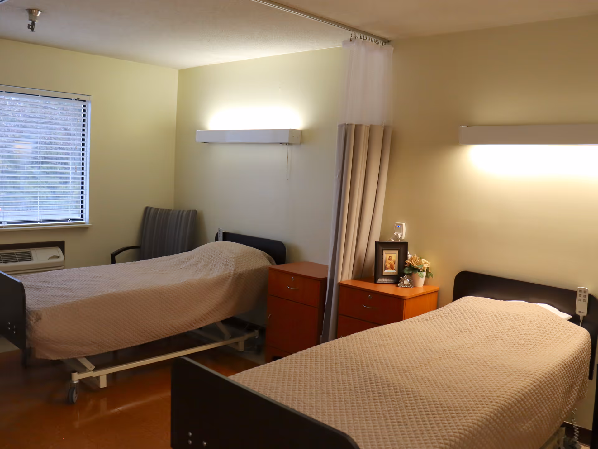 A room with two hospital-style beds covered with beige quilts, separated by a privacy curtain. Each bed has a wooden nightstand with drawers beside it. One nightstand has a framed picture and a small potted plant. There is a window with blinds and a chair near one bed. The walls are light-colored and there are wall-mounted lights above each bed.