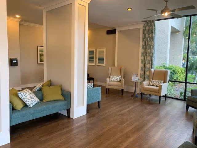 Bright seating area with upholstered chairs and sofas, decorative pillows, wood flooring and a large window overlooking greenery.