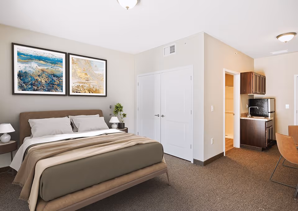 Bright furnished bedroom with a made bed, two nightstands and artwork above the headboard, double closet doors, and a small kitchenette visible through an open doorway.