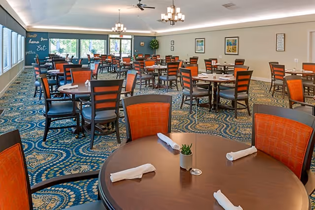 A spacious dining room with multiple round tables and chairs featuring orange backs and dark frames. Each table is set with white rolled napkins and a small plant centerpiece. The room has large windows letting in natural light, patterned carpet, and modern ceiling lights.