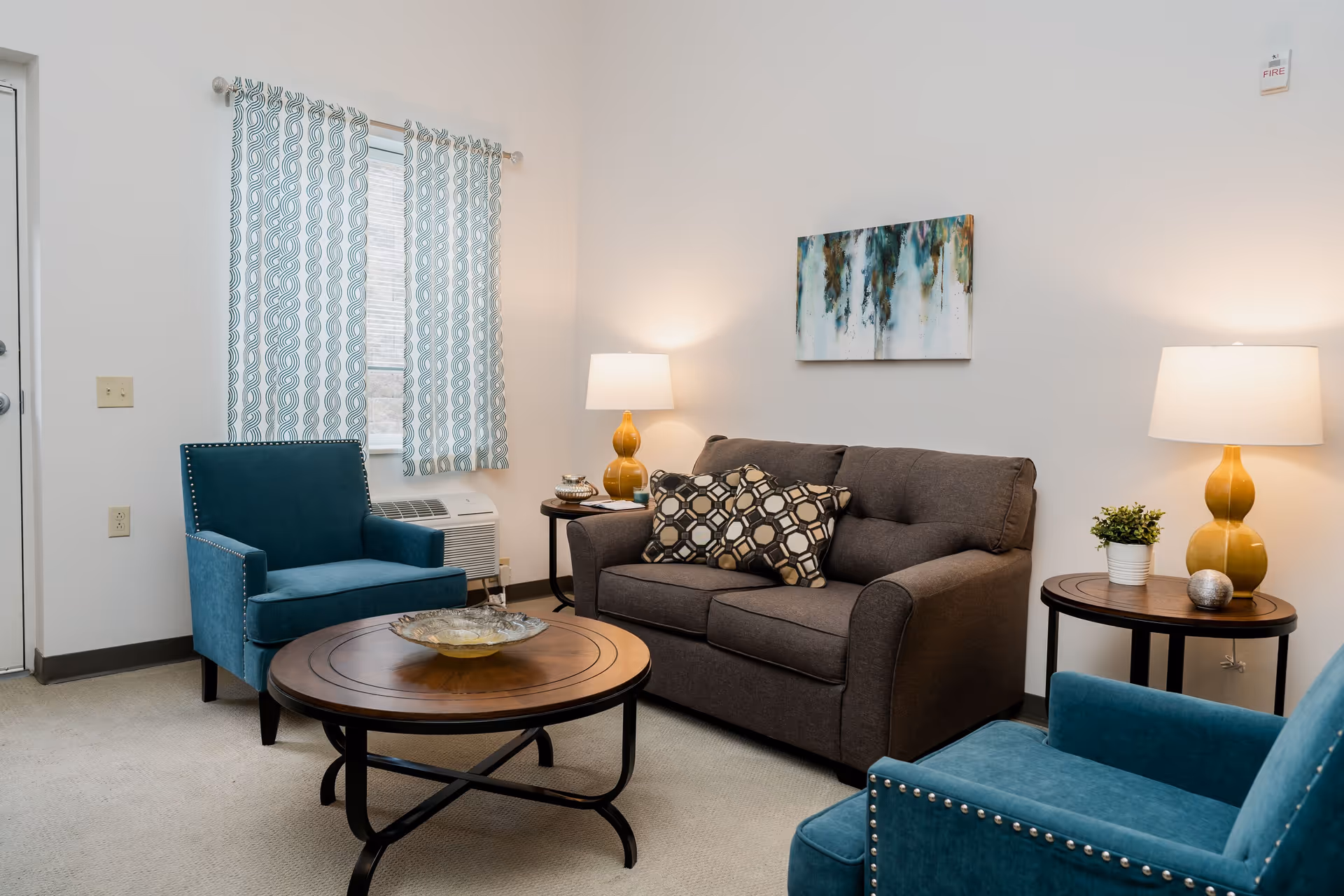 Well-lit living room with a gray loveseat, two teal armchairs, a round wooden coffee table, side tables with lamps, and wall art.