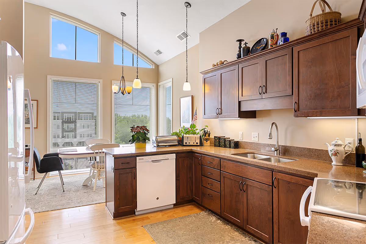 Bright kitchen area with wooden cabinets, a double sink, a dishwasher, and a countertop with various kitchen items. In the background, there is a dining area with a table, chairs, large windows, and a high ceiling with pendant lights.