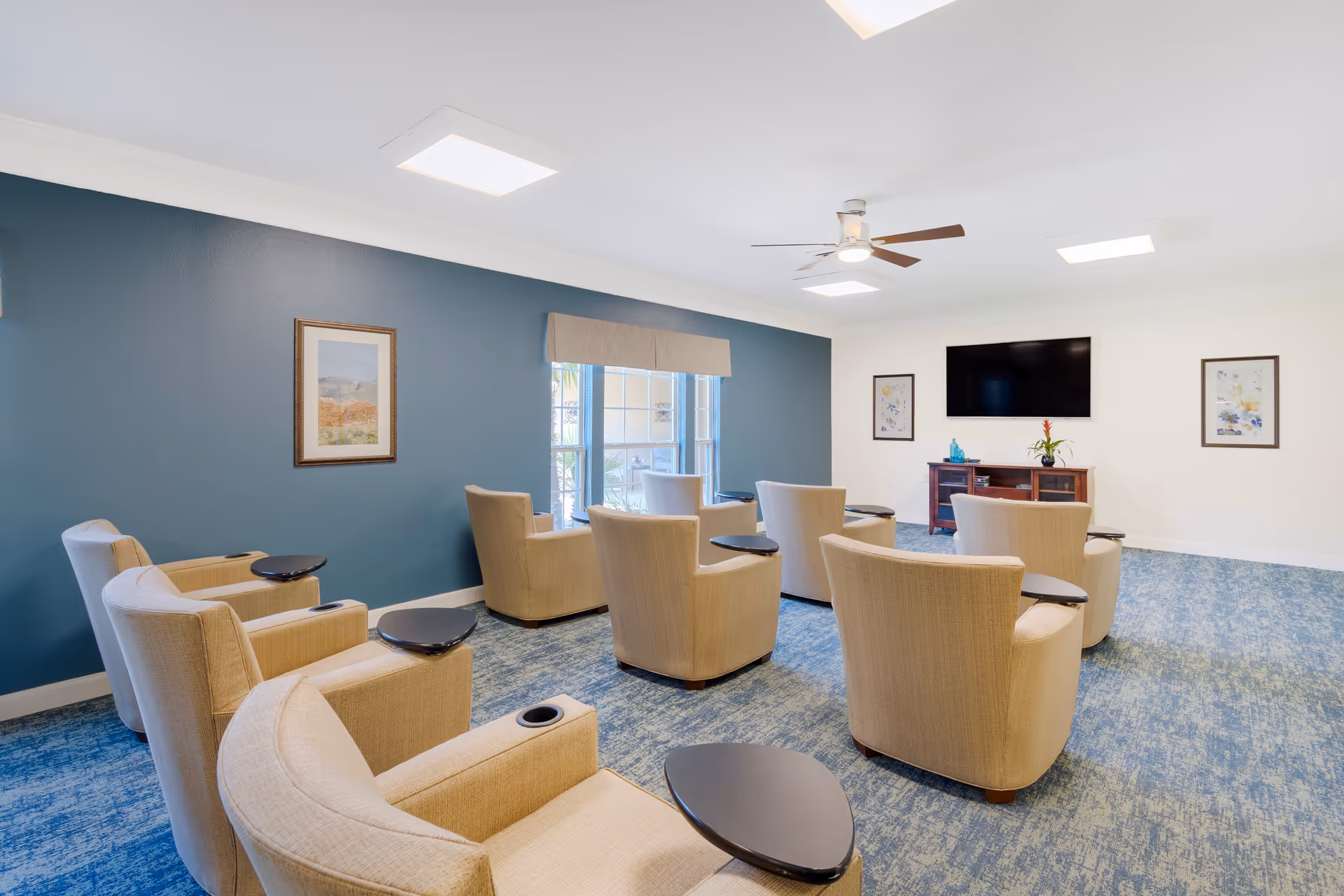 A bright and spacious living room with beige armchairs arranged in rows facing a wall-mounted flat screen TV. The room features a blue accent wall with a framed picture, large windows with beige valances, a ceiling fan with light, and blue patterned carpet. There are two framed pictures on the white wall flanking the TV, and a wooden media console below the TV with decorative items.