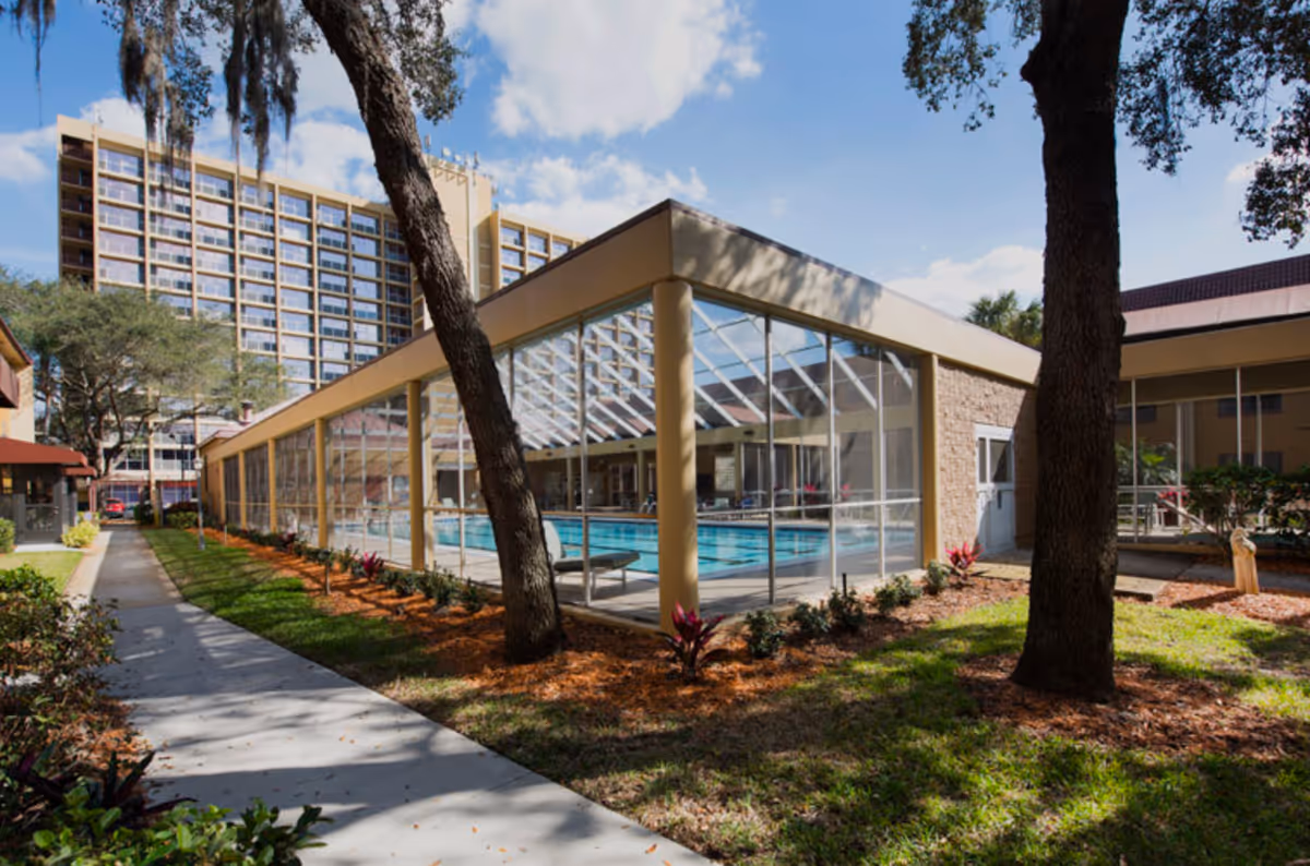Outdoor view of a senior living facility showing a glass-enclosed indoor swimming pool with lounge chairs inside. The building has beige walls and large windows, surrounded by landscaped grass, trees, and plants. A tall multi-story building is visible in the background under a partly cloudy sky.