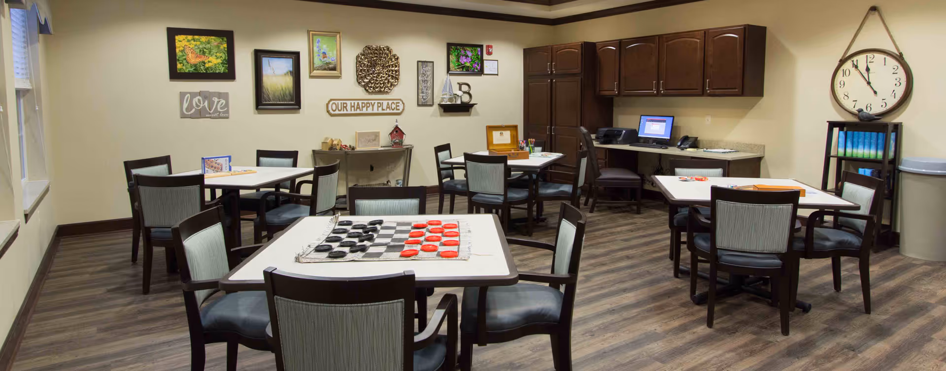 A cozy common room with several tables and chairs arranged for group activities. One table has a checkers game set up. The walls are decorated with framed pictures, a clock, and signs including one that says 'OUR HAPPY PLACE'. There is a desk area with a computer and office supplies in the corner.
