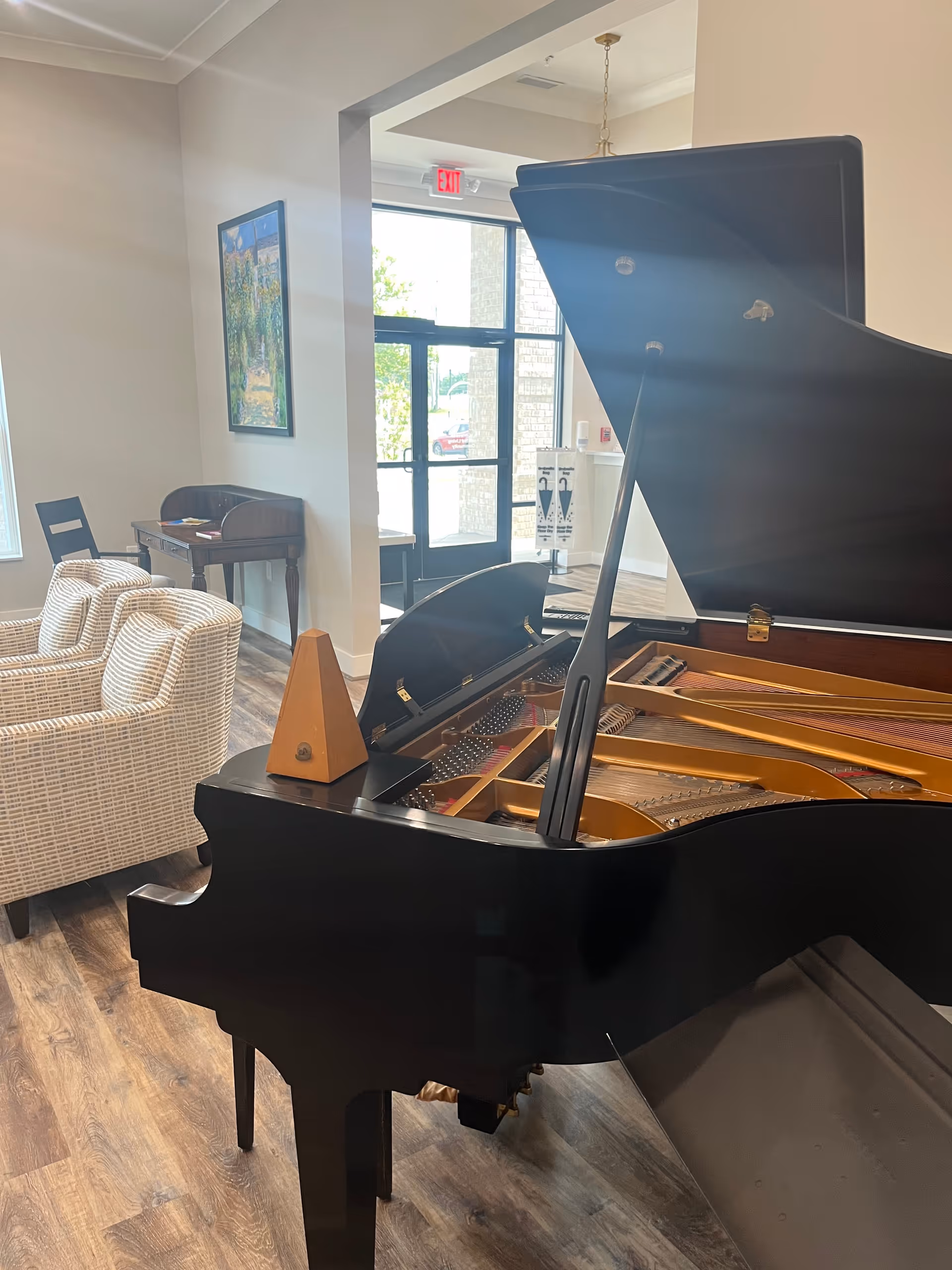 Interior view of a senior living facility lounge area featuring a black grand piano with its lid open, a wooden metronome on top, two beige patterned armchairs, a small wooden desk with a chair, a framed painting on the wall, and a glass door leading outside with an exit sign above it.